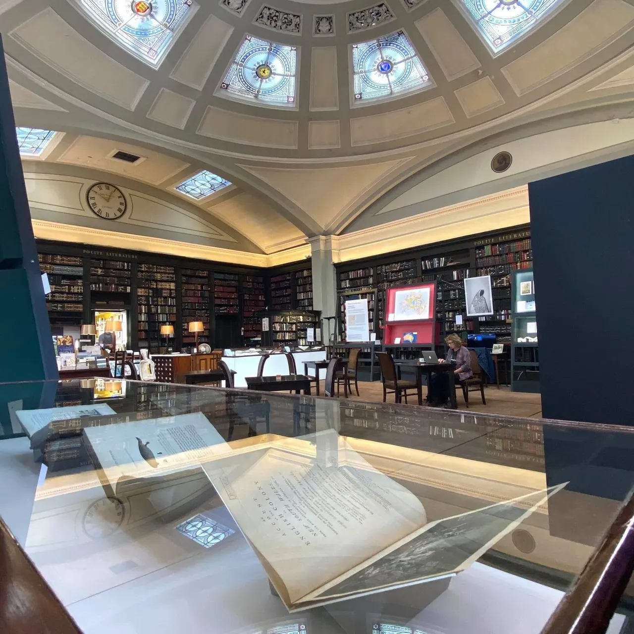 Historic library interior with ornate domed ceiling, illuminated stained-glass skylights, floor-to-ceiling bookshelves, and open manuscripts displayed on tables.