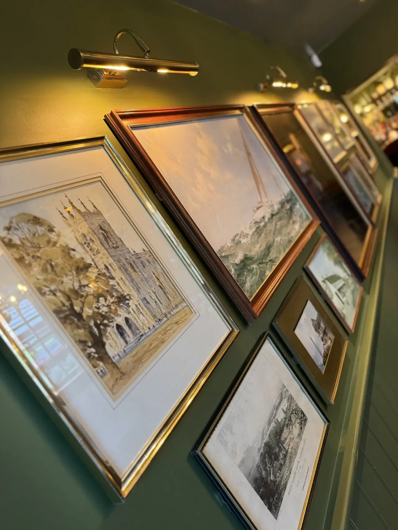 Framed prints and engravings of architectural landmarks and landscapes hang on a green wall, illuminated by brass picture lights.