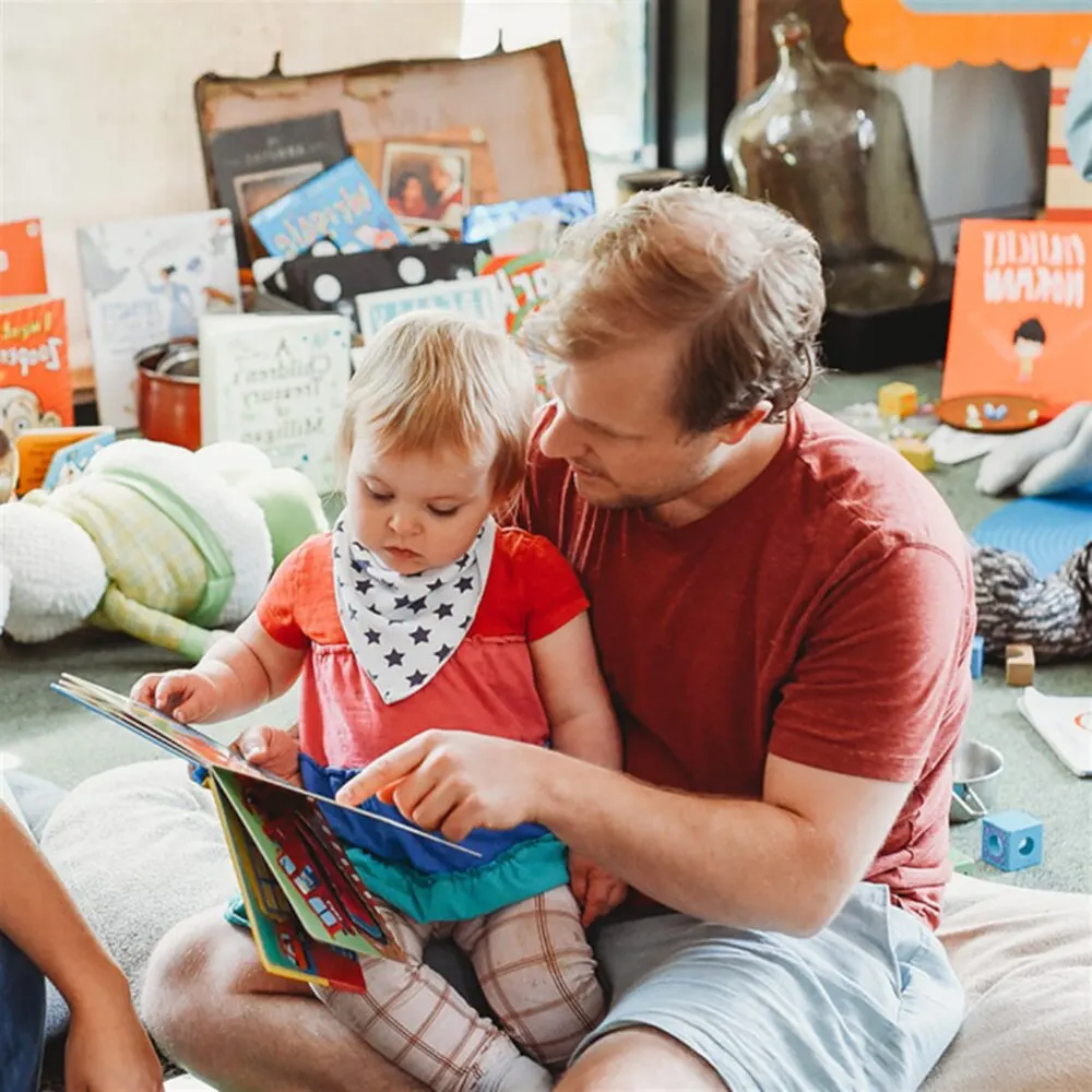 Adult and toddler reading a picture book together on the floor surrounded by toys and children's items.