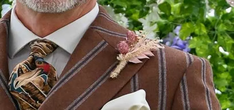 Man in brown tweed jacket wearing a pheasant brooch and holding dried flowers at a racecourse event.