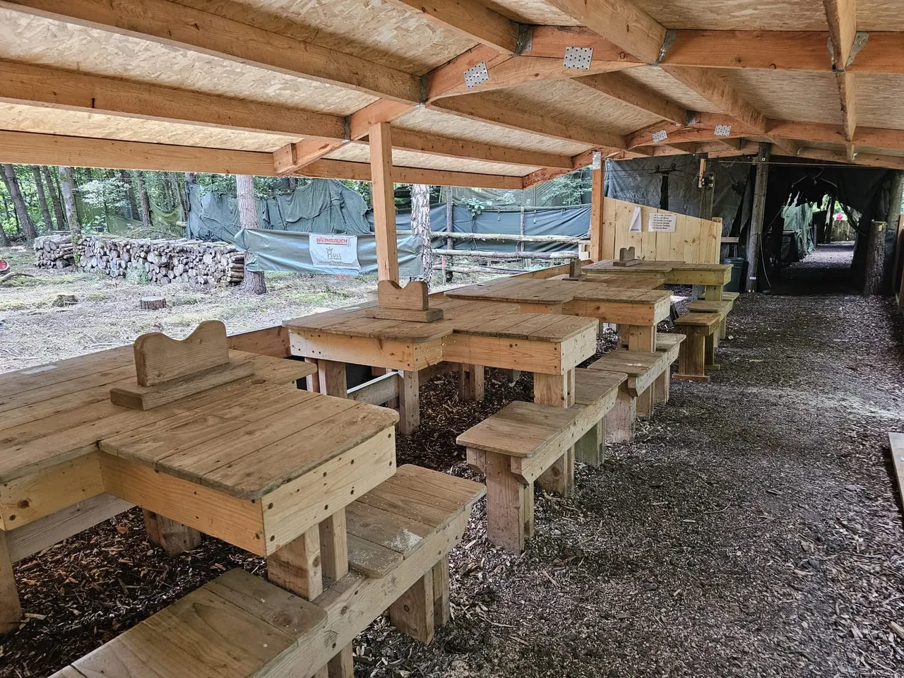 Covered wooden picnic tables and benches in a forest clearing, with log piles and farm enclosures visible in the background.
