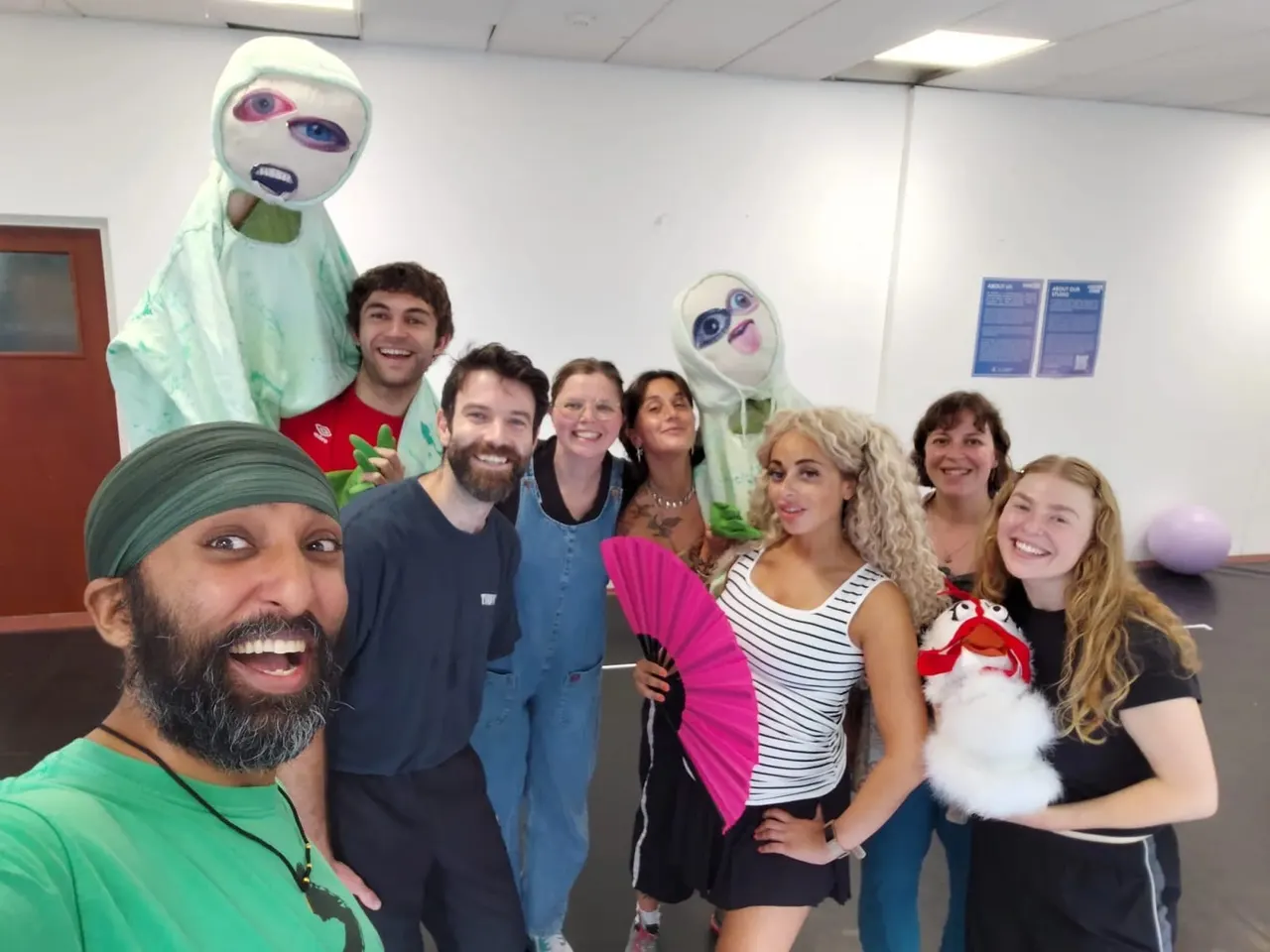 Group of eight adults smiling at camera with colourful face paint and costume masks in an indoor studio space.