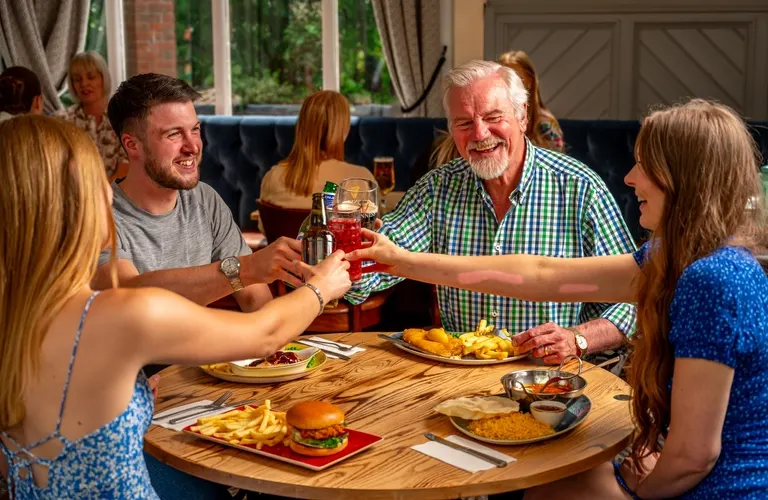 Family and friends gather around a wooden table indoors, clinking glasses and sharing a meal with burgers and chips.