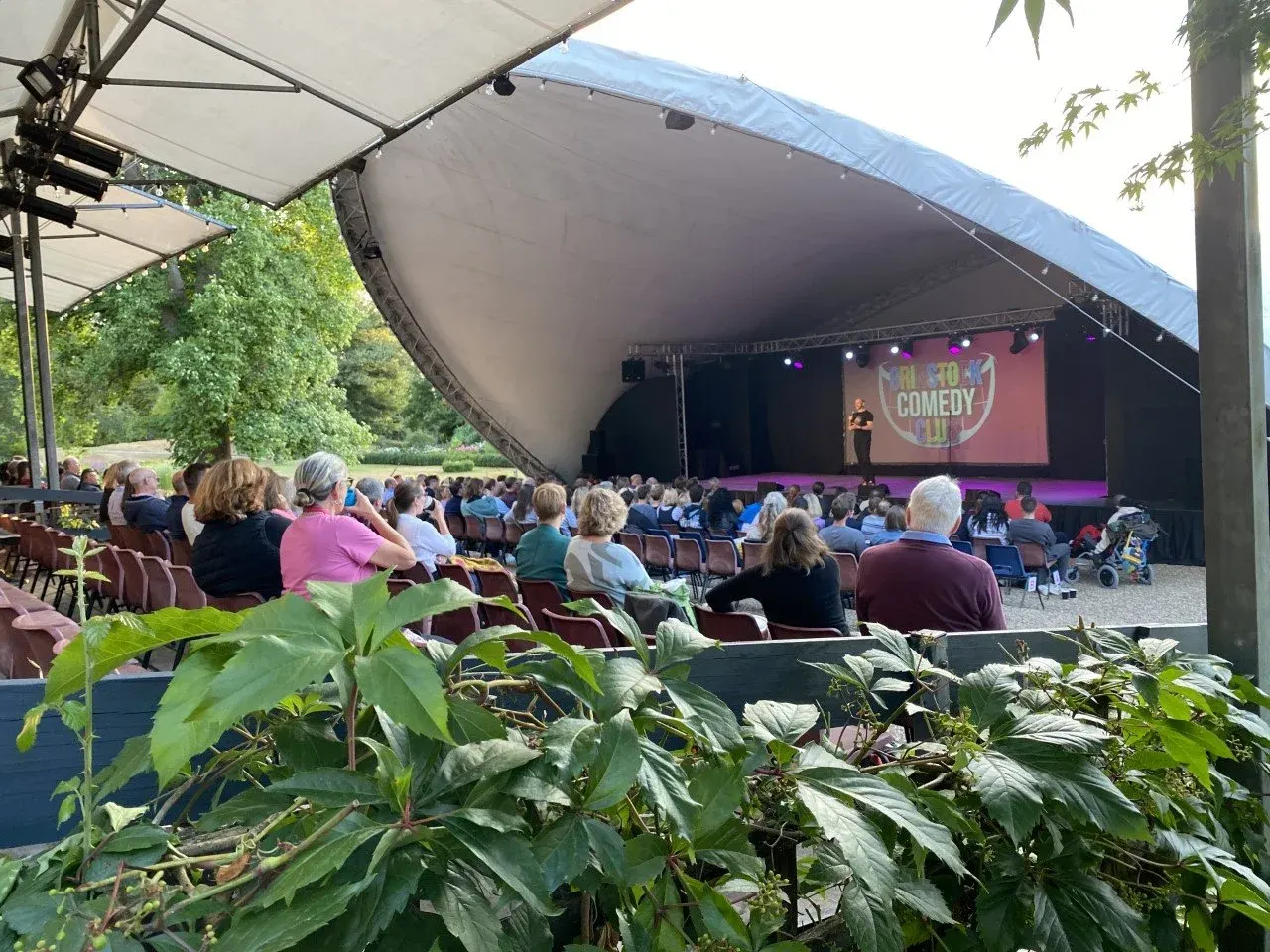 Outdoor comedy festival audience watching a performer on an illuminated stage beneath a large tent canopy.