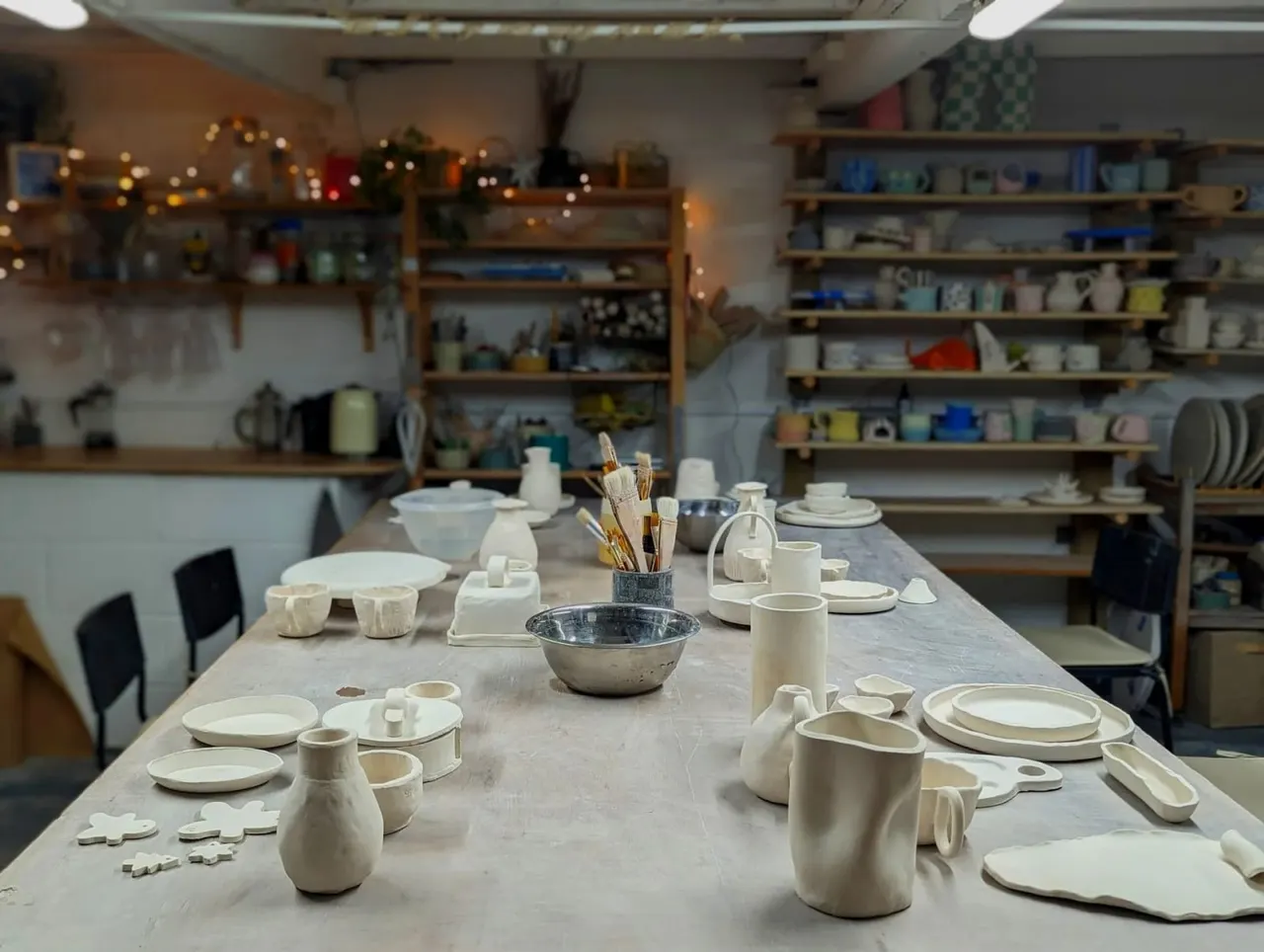 A pottery studio workshop table displaying handmade ceramic pieces including bowls, plates, cups and jugs, with shelves of finished work visible in the background.
