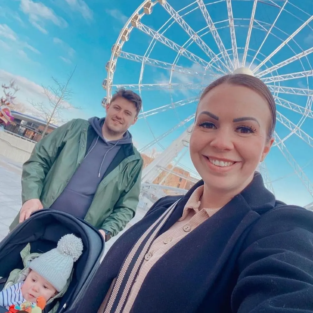 A woman with a baby in a pushchair and a man smile at the camera in front of a large Ferris wheel against a blue sky.