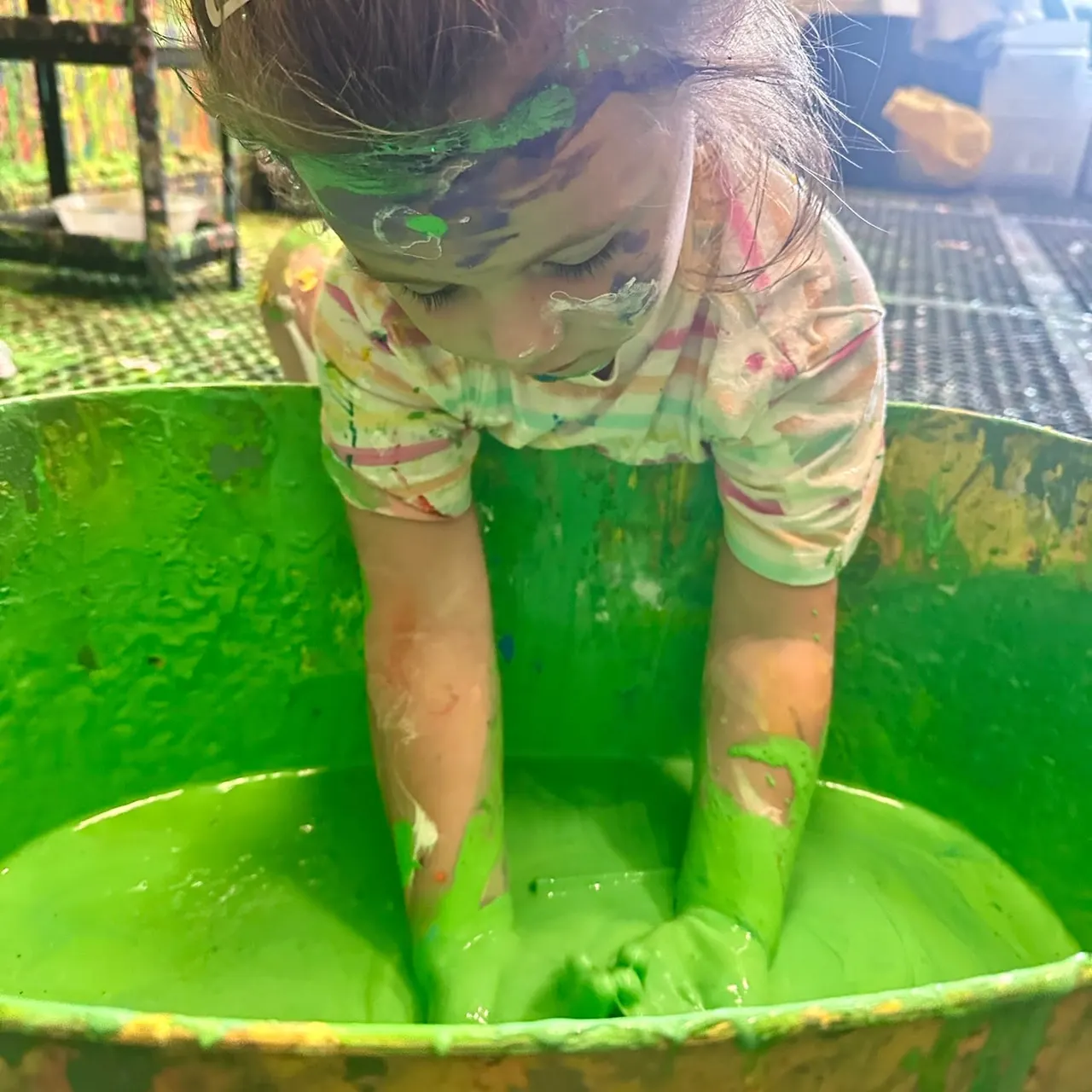 Child covered in green paint playing in a large container of green paint.