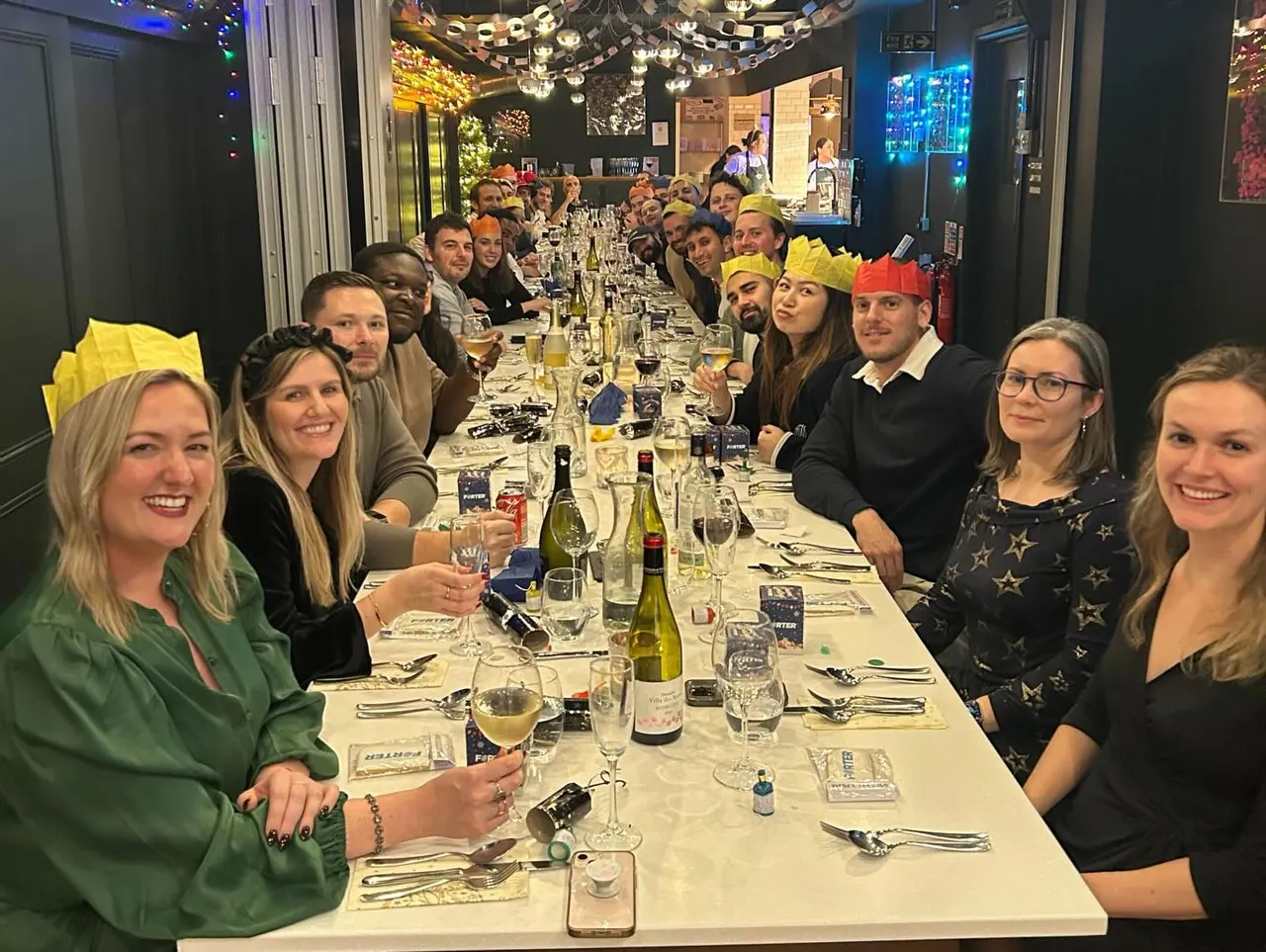 A large group of people seated at a long dining table for a festive meal, many wearing yellow party hats, with wine glasses and place settings visible.