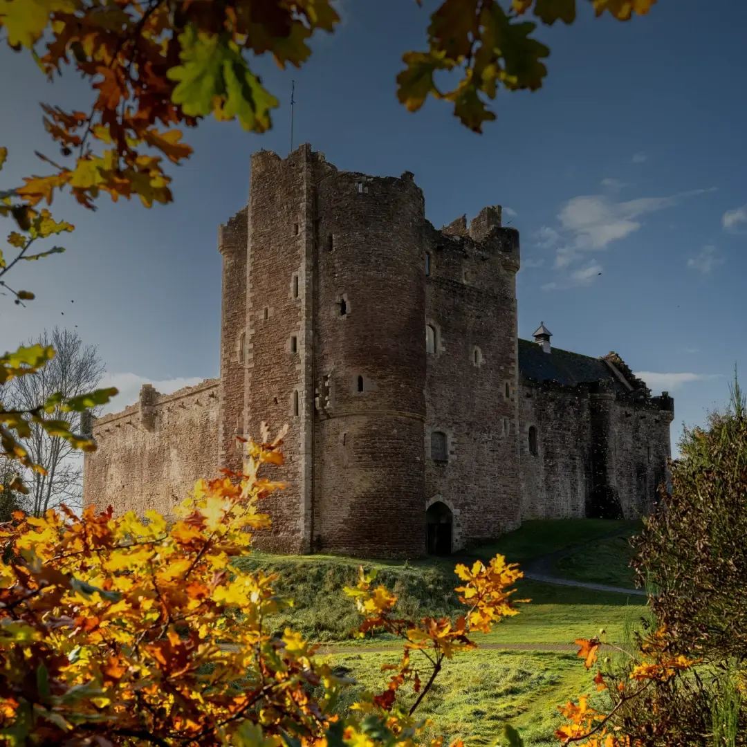 Medieval stone castle with towers viewed across autumn foliage and green grounds under blue sky.