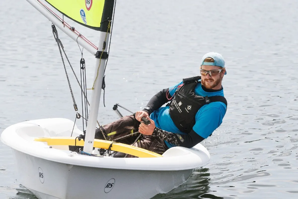 Man in blue and black sailing gear steers a small white sailing boat with yellow sail on calm water.