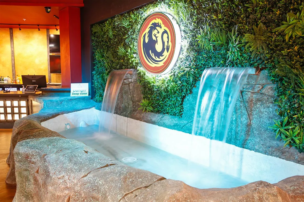Indoor adventure golf course with turquoise pools, cascading waterfalls, neon dragon logo, and tropical vegetation.