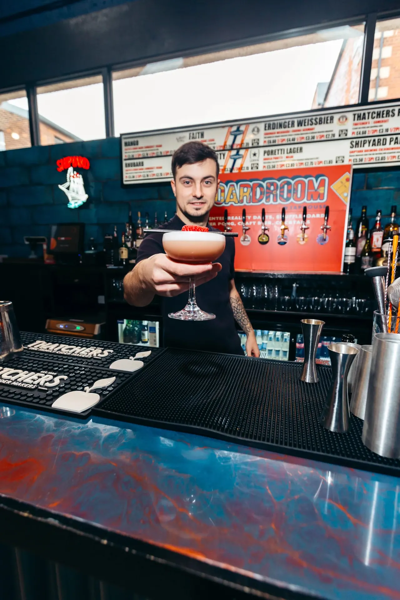 Bartender presenting a cocktail at a bar counter with beer pumps and bottles visible behind.