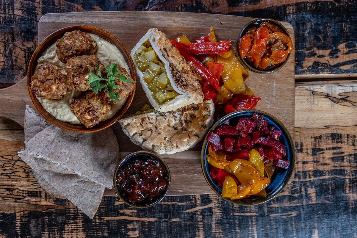 Overhead view of a wooden board with meatballs in sauce, wraps filled with shredded meat and pickled vegetables, and bowls of roasted peppers and relish.