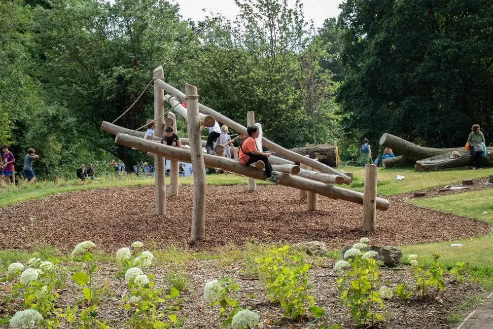 Children climbing and playing on a large wooden log structure in a natural play area surrounded by trees.