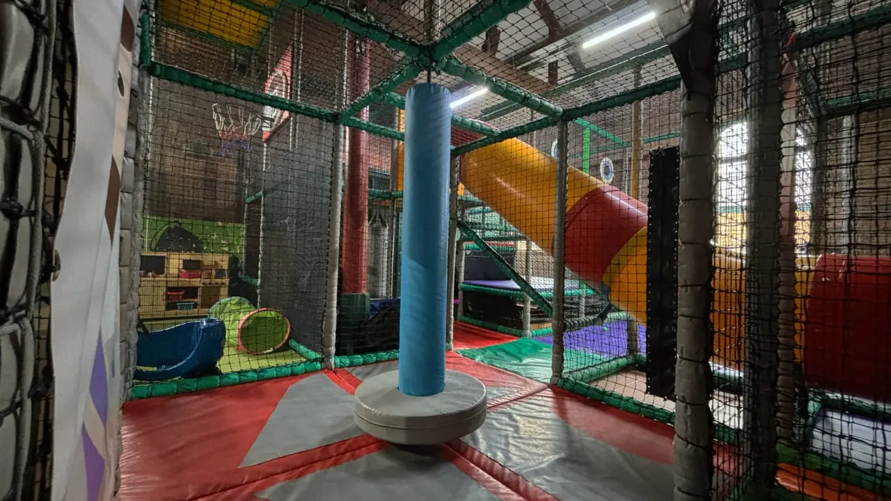 Interior of an indoor play centre with colourful soft play equipment, slides, and netting enclosed within a green metal frame structure.