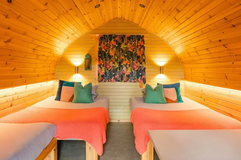 Interior of a wooden glamping pod with two beds with orange blankets and green cushions, warm lighting, and floral artwork.