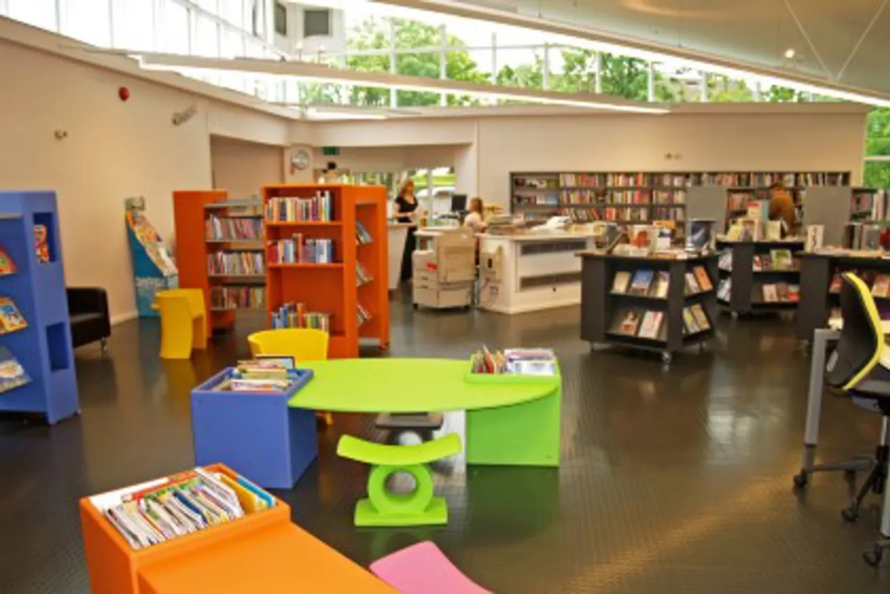 Modern library interior with colourful shelving units, reading tables and book displays on dark flooring, staff visible at desk in background.