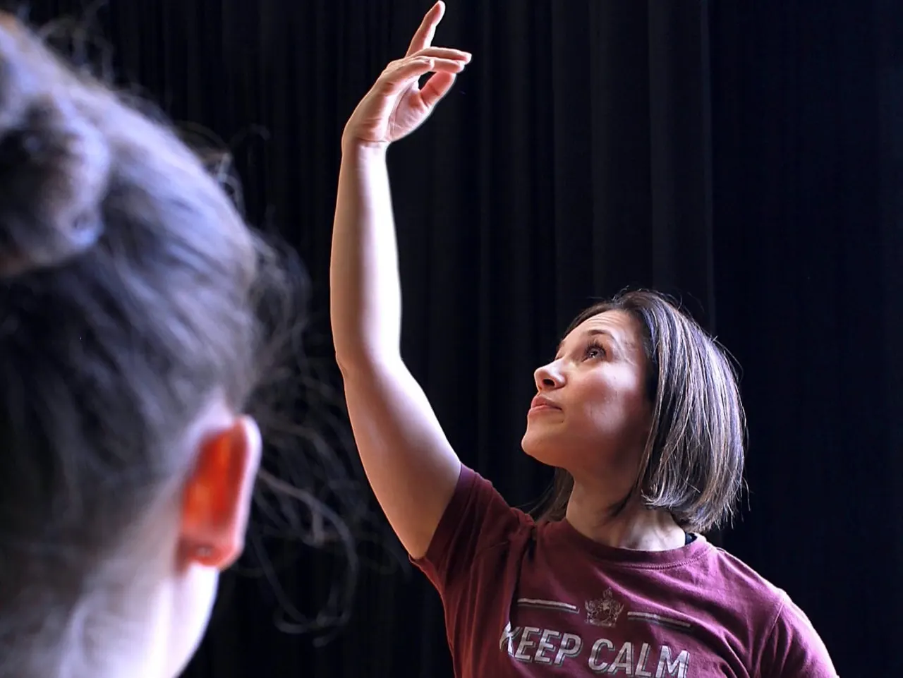 A woman in a burgundy t-shirt raises her arm whilst dancing with a partner against a dark curtained background.