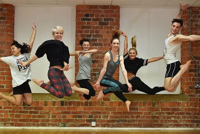 Group of six people jumping and posing mid-air in a studio with exposed brick walls.
