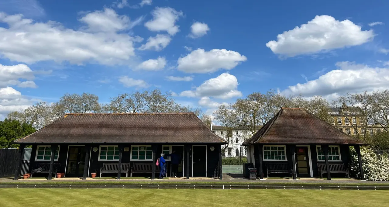Black pavilion buildings with pitched roofs beside a bowling green, with person in blue jacket nearby on a sunny day.