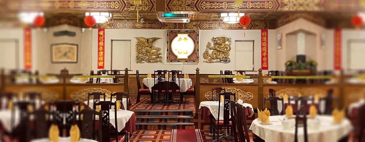 Ornate Chinese restaurant dining room with tables set with white tablecloths, decorative gold panels and red lanterns overhead.