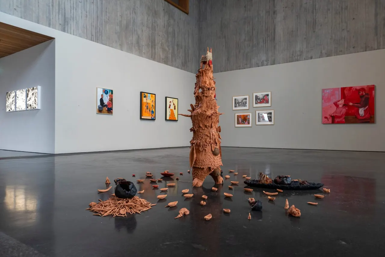 Gallery interior with wooden carved sculptural installation on dark floor surrounded by scattered wooden pieces, and framed artworks displayed on white walls.