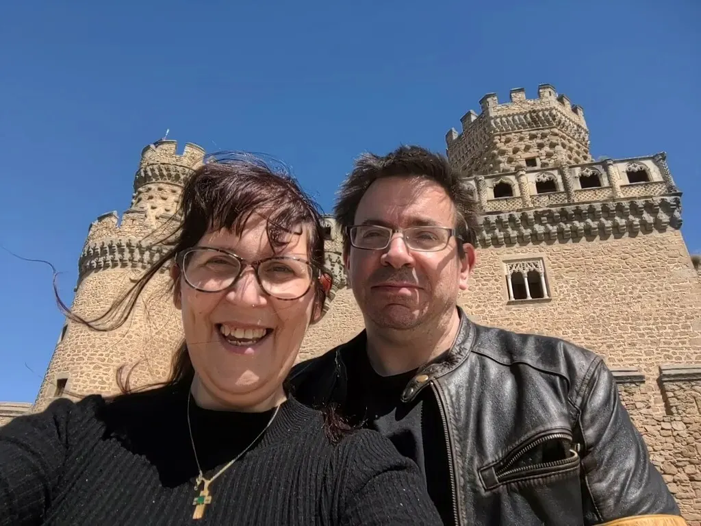 Two people smiling at the camera in front of a historic stone castle with crenellated towers under clear blue sky.