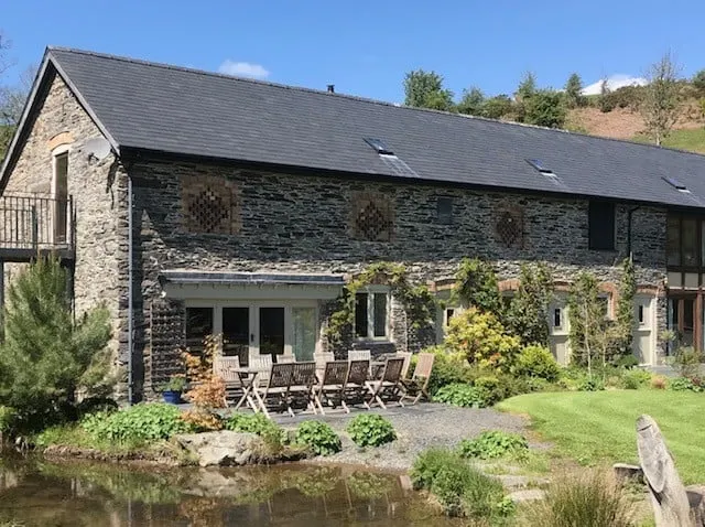 Stone barn conversion with patio seating beside a pond, surrounded by green gardens.