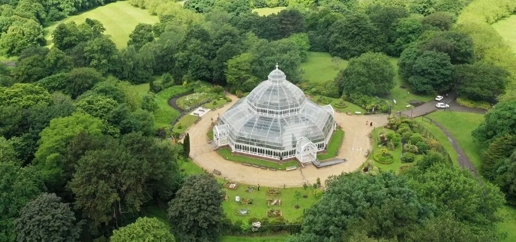 Aerial view of a Victorian glasshouse with domed roof surrounded by tree-filled parkland with visitors on the grounds.