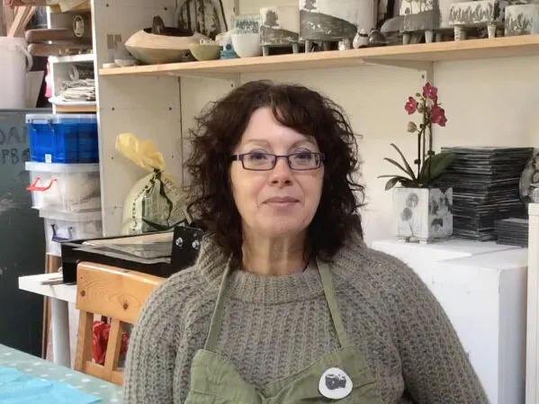 Woman wearing glasses and green apron standing in pottery studio with shelves of ceramics and plants behind her.