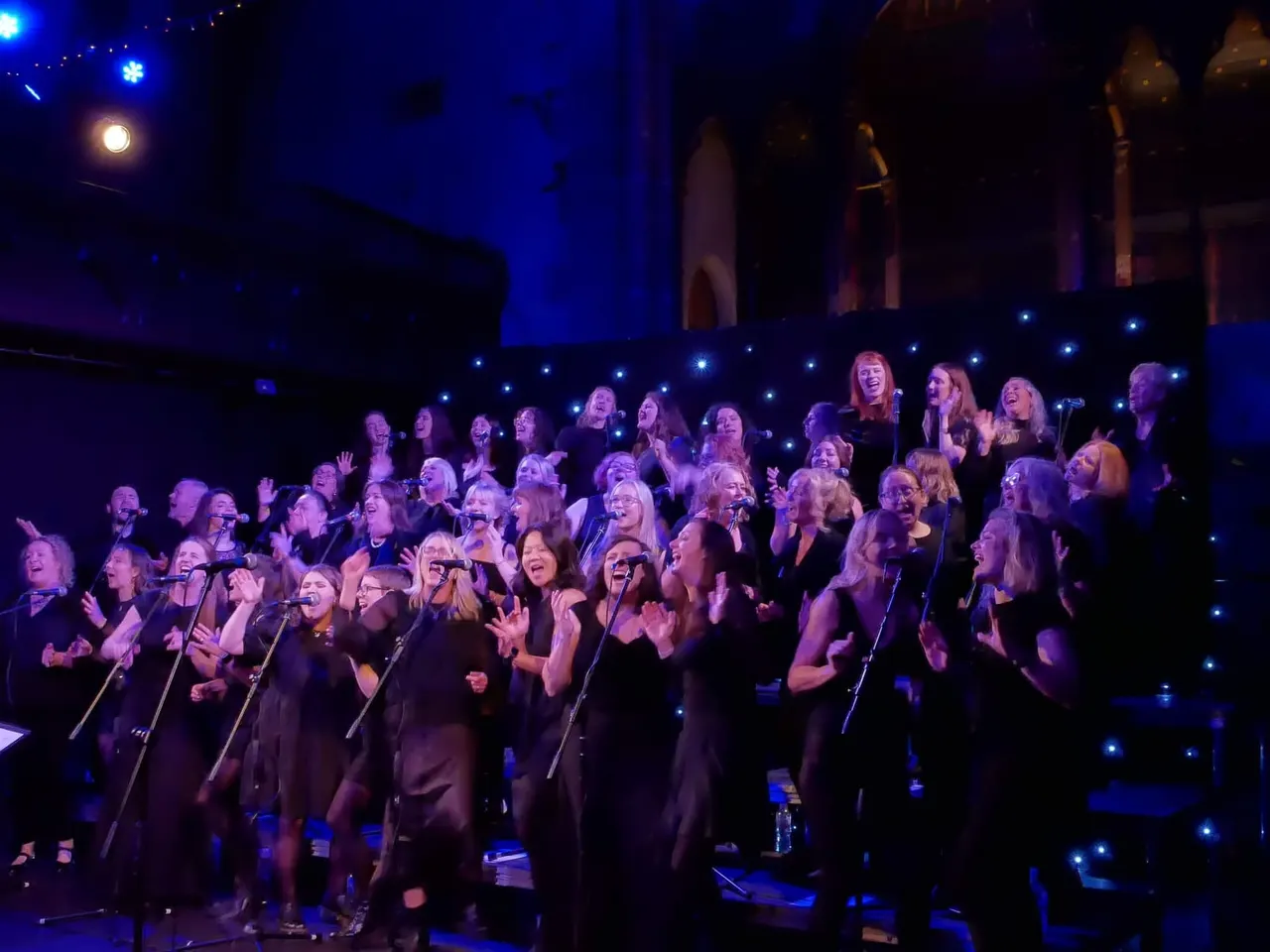 Large choir performing on an illuminated stage with blue lighting and small white lights surrounding the singers.