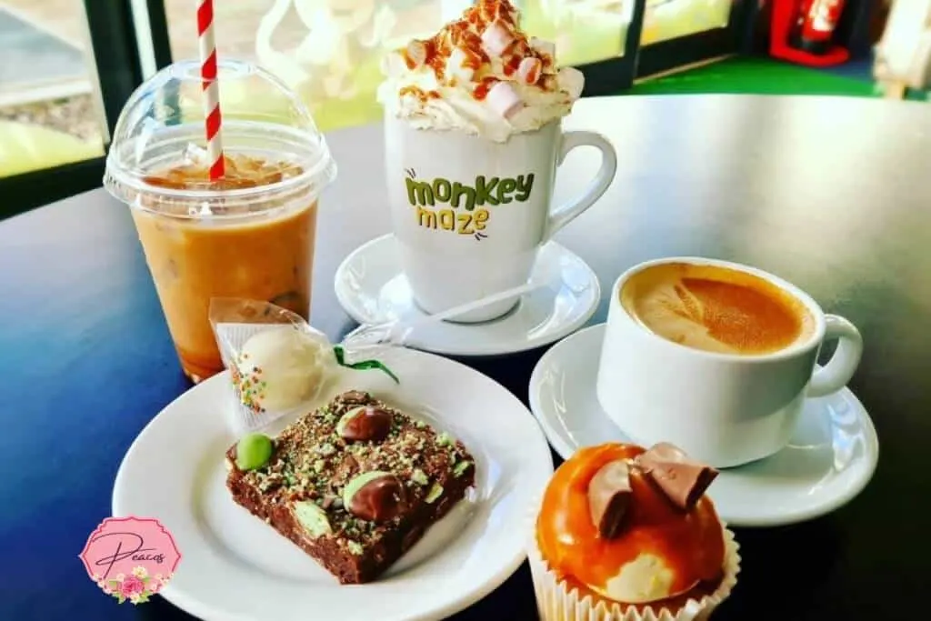 A table spread showing an iced drink with striped straw, a chocolate cake with nuts and berries, two hot coffees, and a topped cupcake.