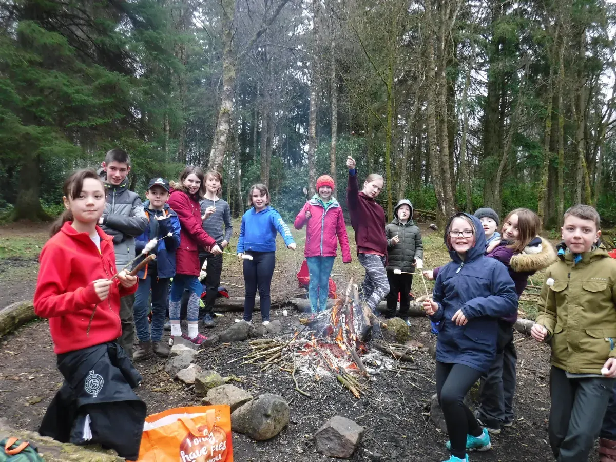 Group of children gathered around a campfire in a woodland clearing, wearing colourful outdoor jackets.