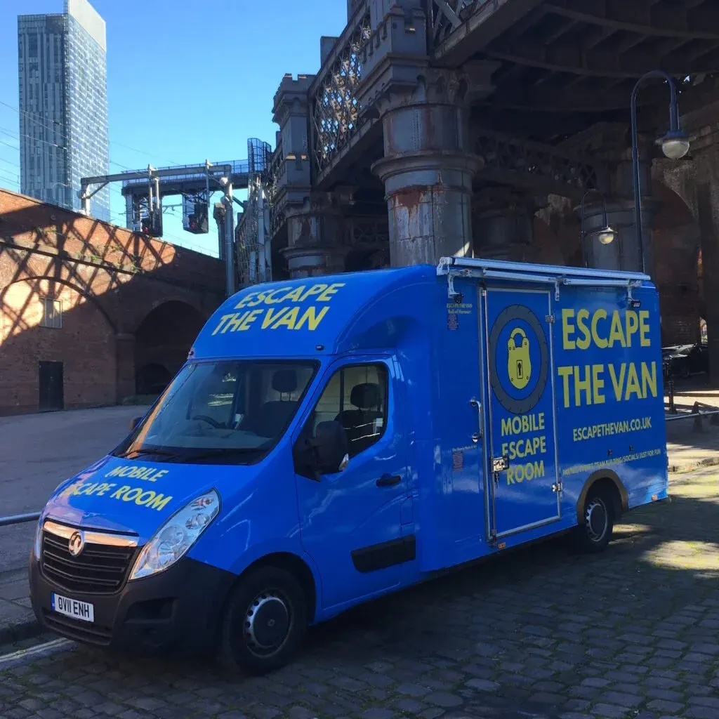 Bright blue mobile escape room van parked in an urban courtyard with historic industrial buildings and modern skyscrapers behind it.