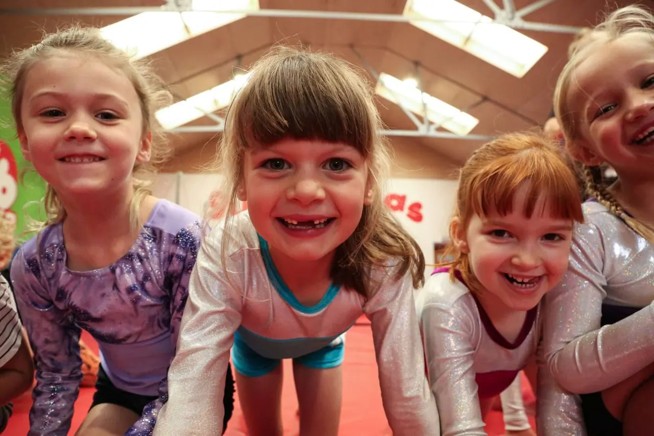 Small children in sparkly leotards smiling at camera in an indoor gymnastics facility.