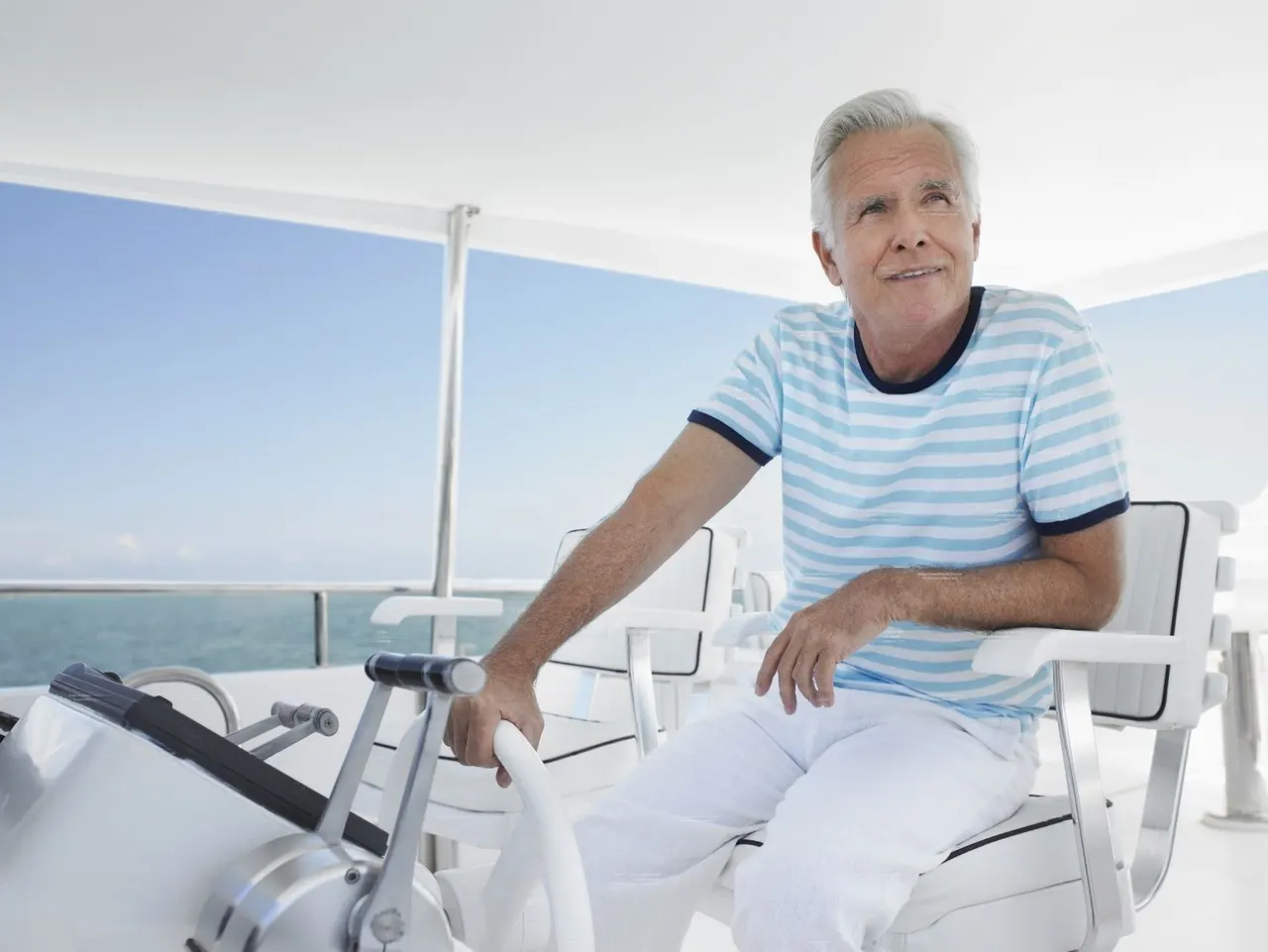 Older man at the helm of a boat on open water, wearing striped blue and white shirt.