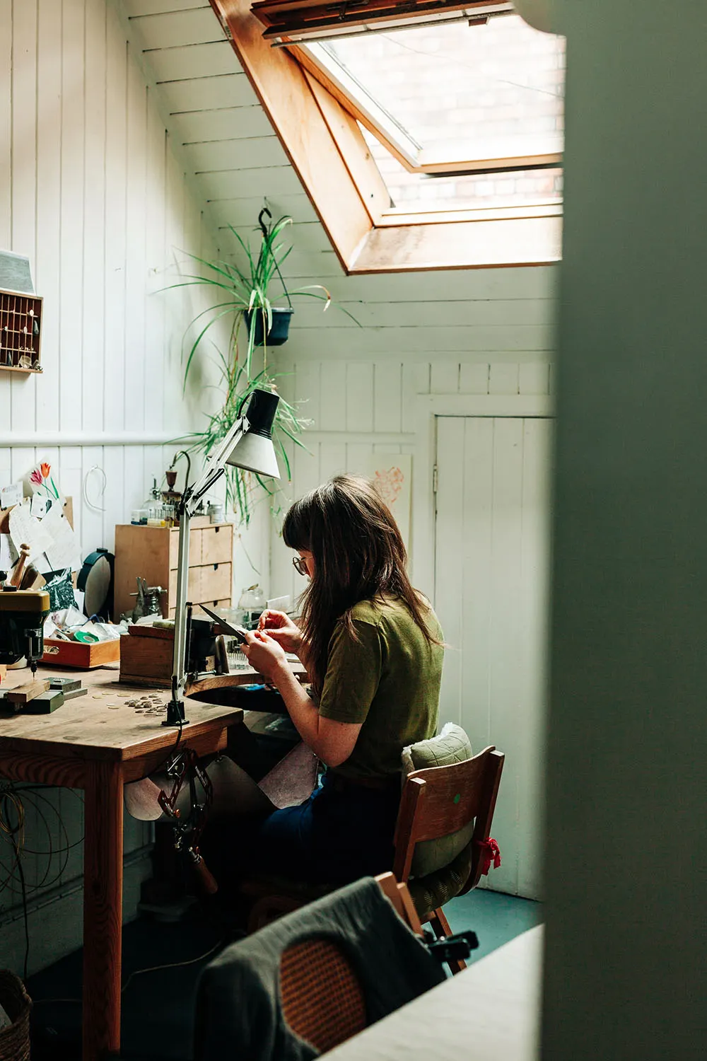 Person working at a wooden desk in a bright studio space with a skylight and potted plants.