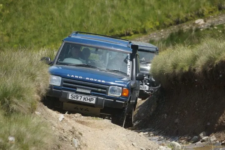 Blue Land Rover navigating a steep, narrow off-road track through grassland and moorland terrain.