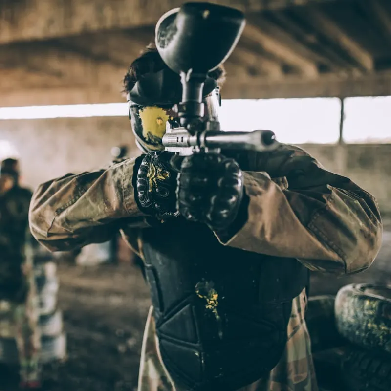 Player in protective gear and helmet aiming paintball marker inside an indoor arena.