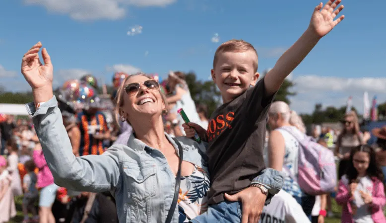 Woman and boy with raised arms celebrating at outdoor event with crowds and fairground rides in background.