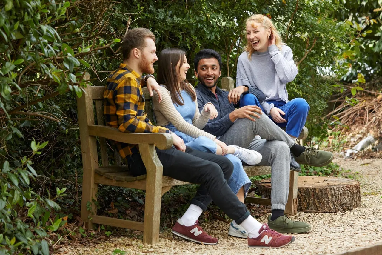 Four young people sitting together on a wooden bench in a leafy garden setting, smiling and chatting.