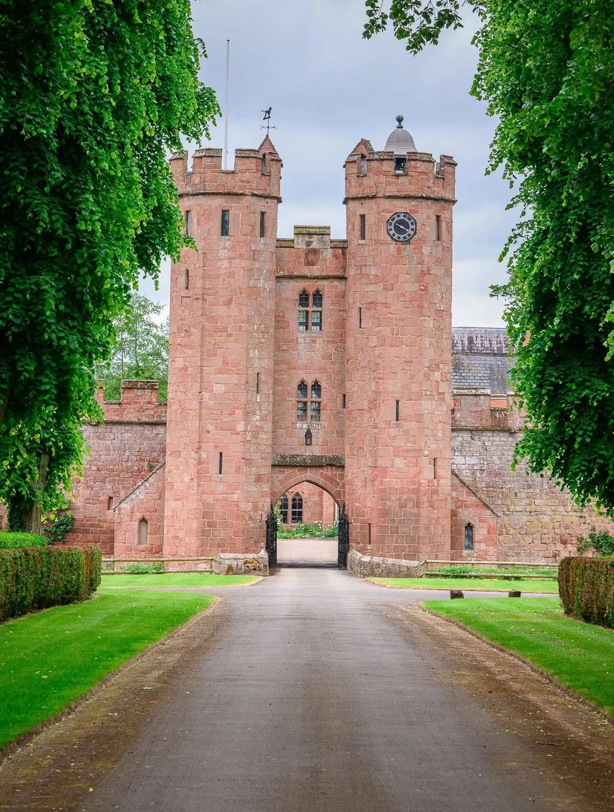 Red sandstone castle with twin towers and clock face, approached via tree-lined driveway with manicured lawns on either side.