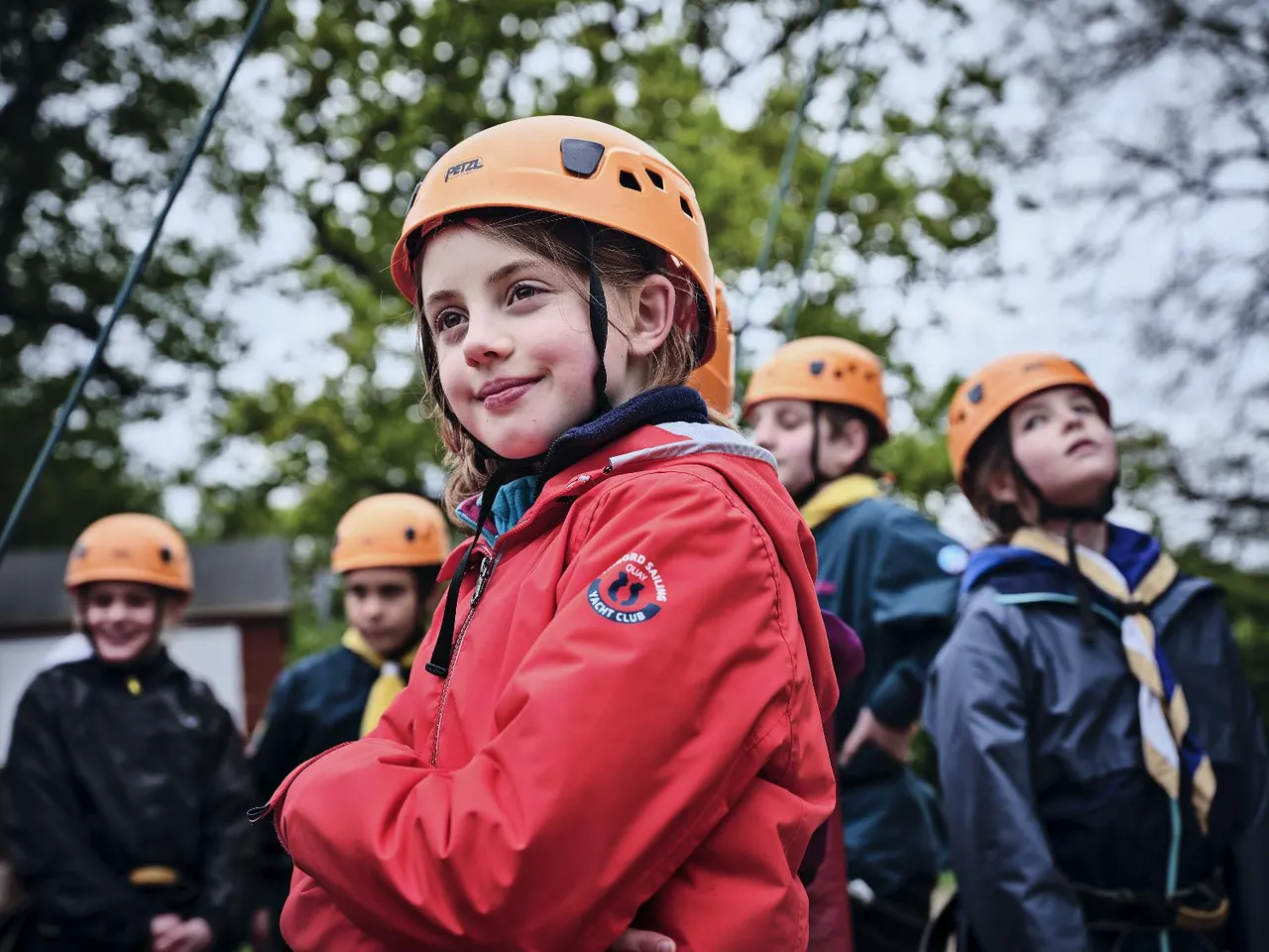 Children wearing orange safety helmets and scout uniforms at an outdoor activity centre surrounded by trees.