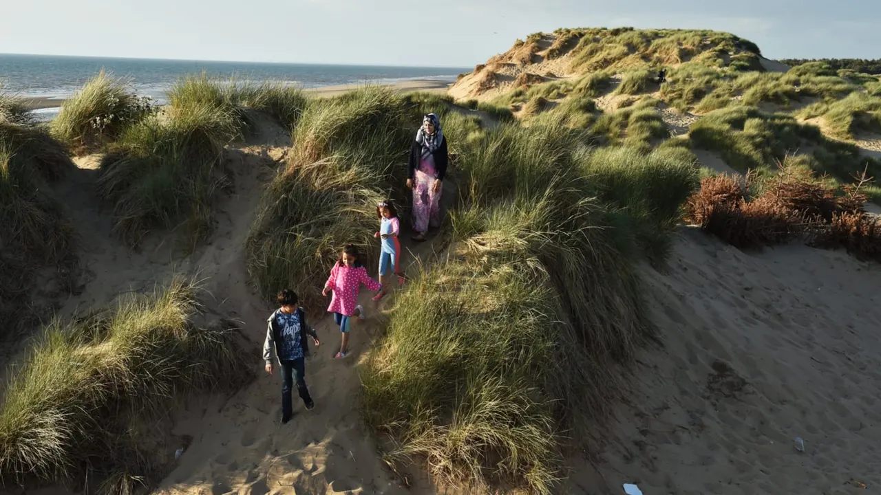 An adult and three children walking through grassy sand dunes towards a beach, with cliffs visible in the distance.