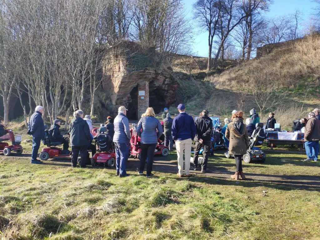 Visitors gather outdoors with red ride-on mowers near a stone cave entrance set into a hillside with bare trees.