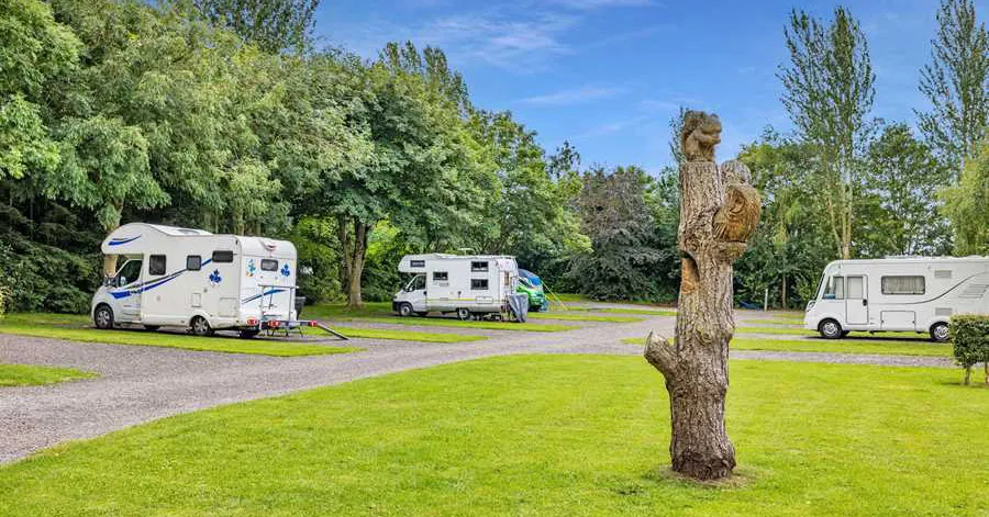 Motorhomes parked on grassy pitches amongst trees at a caravan park, with an old wooden post in the foreground.