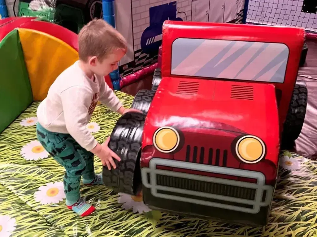 A young child playing with a large red toy car in an indoor play area with colourful soft play equipment.