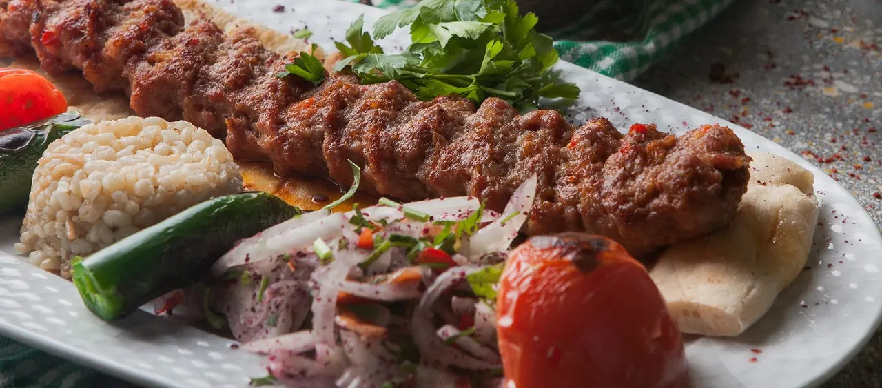 Grilled meat kebabs served with rice, salad and roasted vegetables on a white plate.