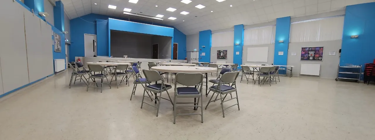 Large modern hall with blue pillars and white walls, set up with multiple round tables and grey chairs, a stage at far end, and bright ceiling lights.