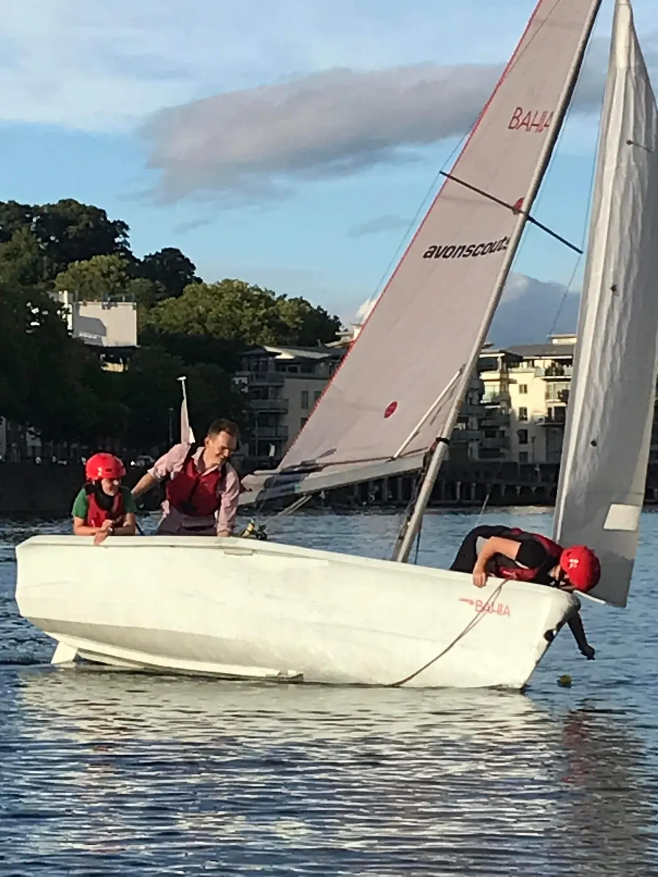 Three young people in red helmets sailing a small dinghy on calm water near waterfront buildings.